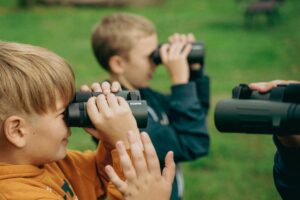 Two boys using binoculars to observe nature closely on a grassy outdoor setting.